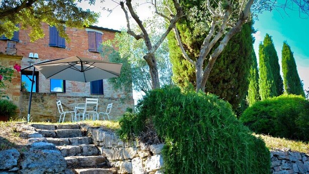 Una escalera de piedra conduce a un área de estar al aire libre con una mesa y sillas, un paraguas grande y un edificio con ventanas y contraventanas al fondo.