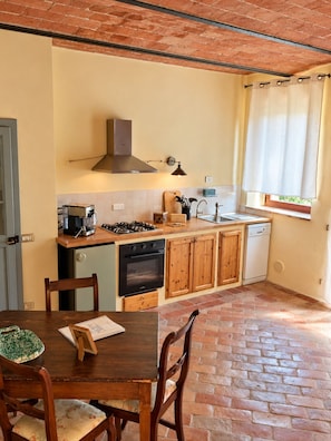 A compact kitchen with a gas stove, oven, and a dishwasher. There is a wooden dining table with chairs, and a window with curtains. - "Il Leccio" Apartment in an authentic Tuscan farmhouse with pool (Toscana)