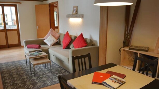 Ein Wohnzimmer mit einer Couch, einem Esstisch mit Stühlen, einem gemusterten Teppich und einer Lampe.