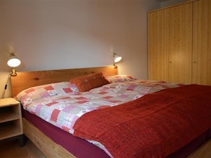 A bed with a wooden headboard, two bedside lamps, a red blanket, and a quilt with a patchwork design. - Oehrli Appartement/Fewo, Dusche oder Bad, WC 4-Bettwohnung by Interhome (Gstaad)