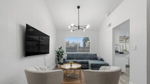 Een moderne woonkamer met een flatscreen televisie, een grijze bank, twee fauteuils, een salontafel en een plafondlamp.