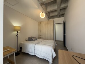 Ein Schlafzimmer mit Bett, Nachttisch, Lampe, Kleiderschrank und einer Tür zu einem anderen Raum.