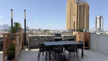 A rooftop terrace with a grill, dining table, chairs, and potted plants.