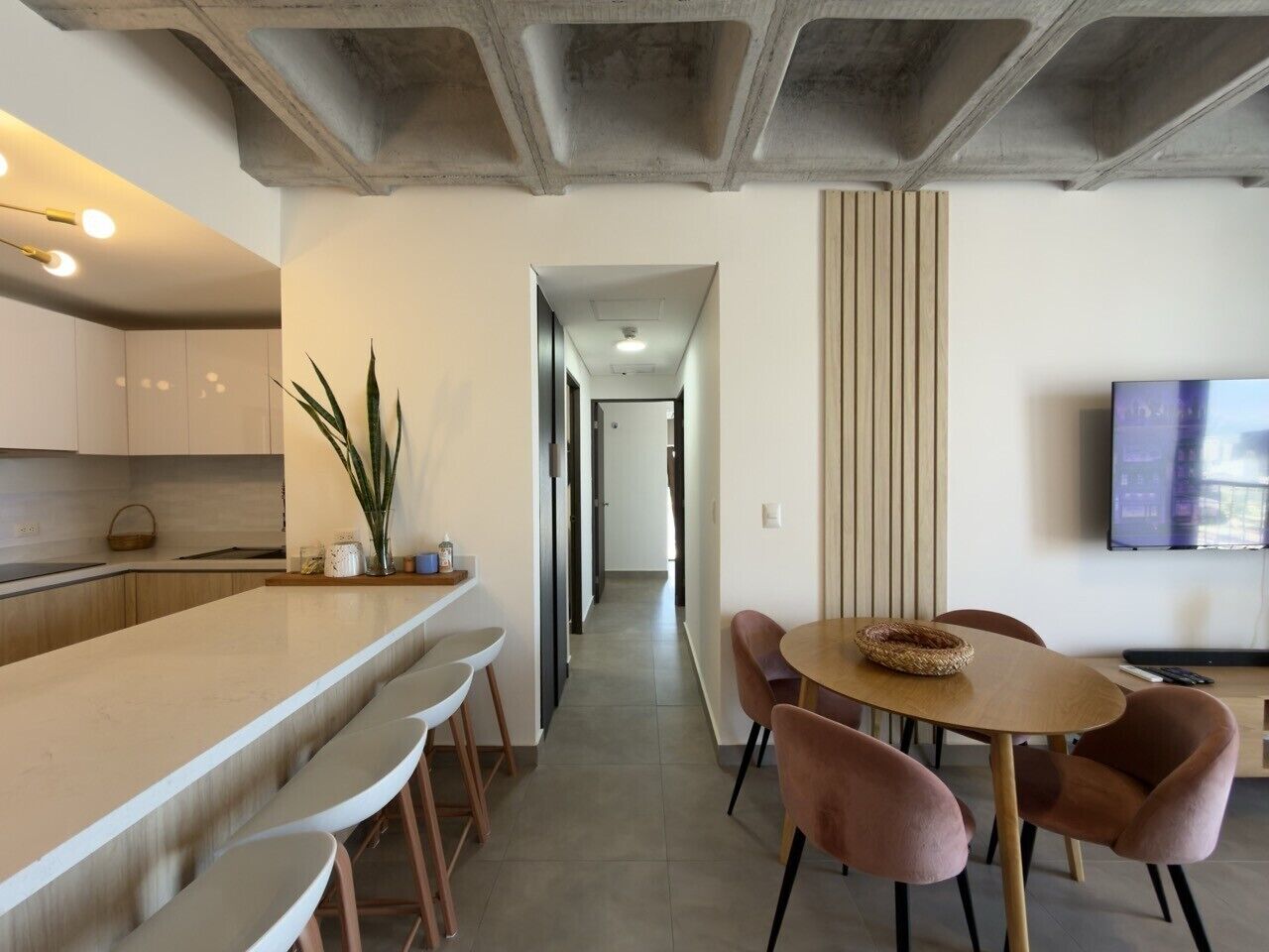 A modern kitchen with a long counter and bar stools, a dining area with a round table and chairs, and a TV mounted on the wall.