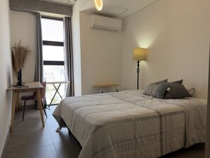 Ein Schlafzimmer mit Bett, Schreibtisch, Stuhl, Lampe und einer Klimaanlage.