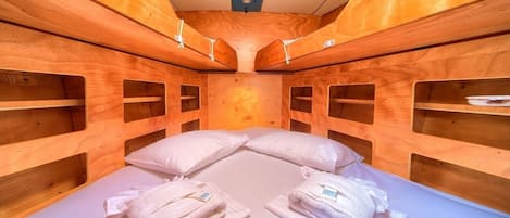 A compact, wooden cabin-style room with bunk beds, built-in shelves, and a central bed with pillows and towels.