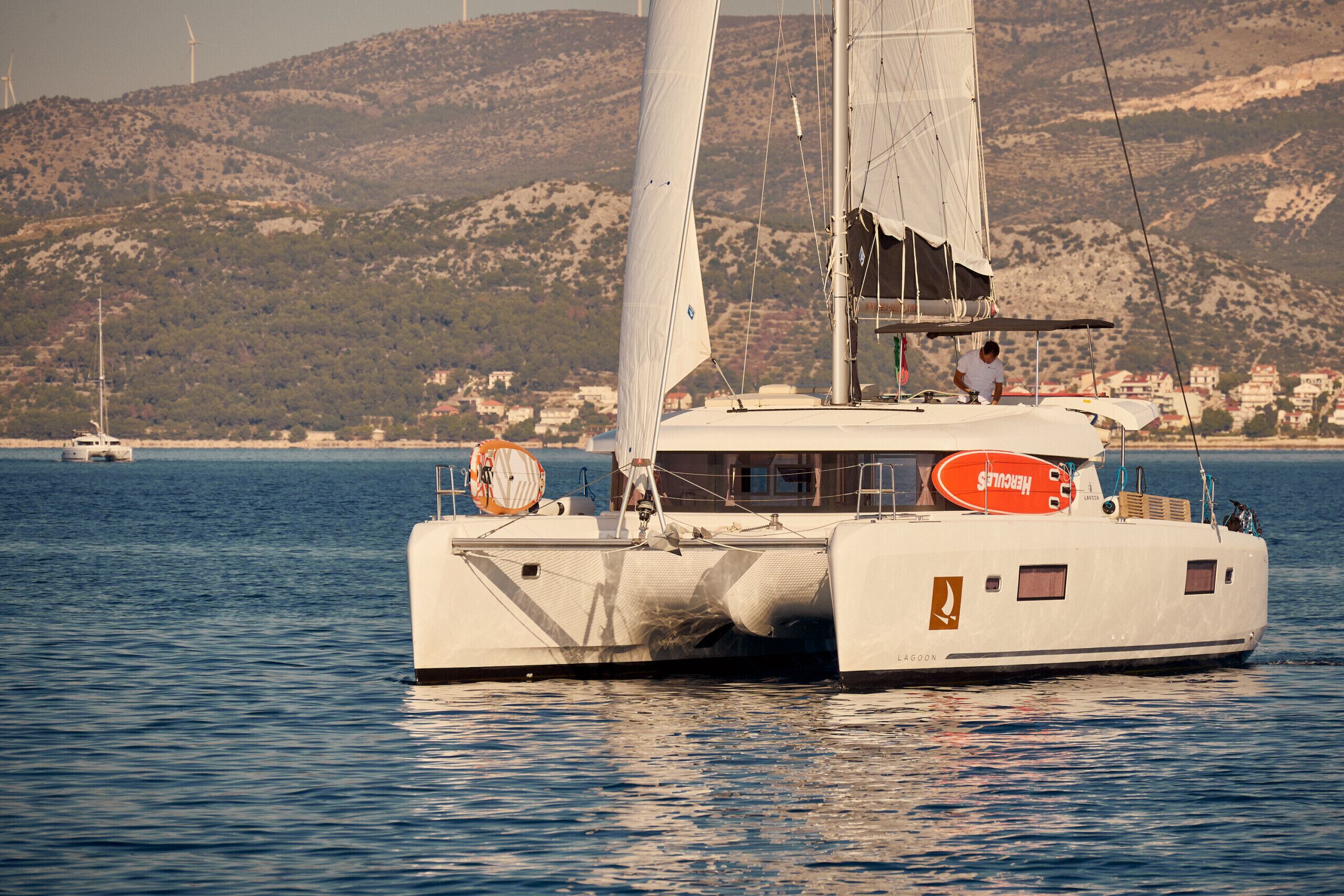 A sailboat with a lifebuoy and a 'Hercules' sign on the deck, sailing on calm waters with a mountainous coastline in the background.