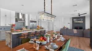 Une cuisine moderne dotée d’un îlot central, un coin repas avec une table en bois mise pour le repas, et un espace de vie avec un canapé et un téléviseur.