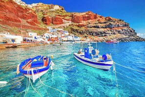Un village côtier où des bateaux sont amarrés dans une eau d’un bleu limpide, des bâtiments blancs rehaussés de touches de bleu, et une colline rocheuse en arrière-plan.