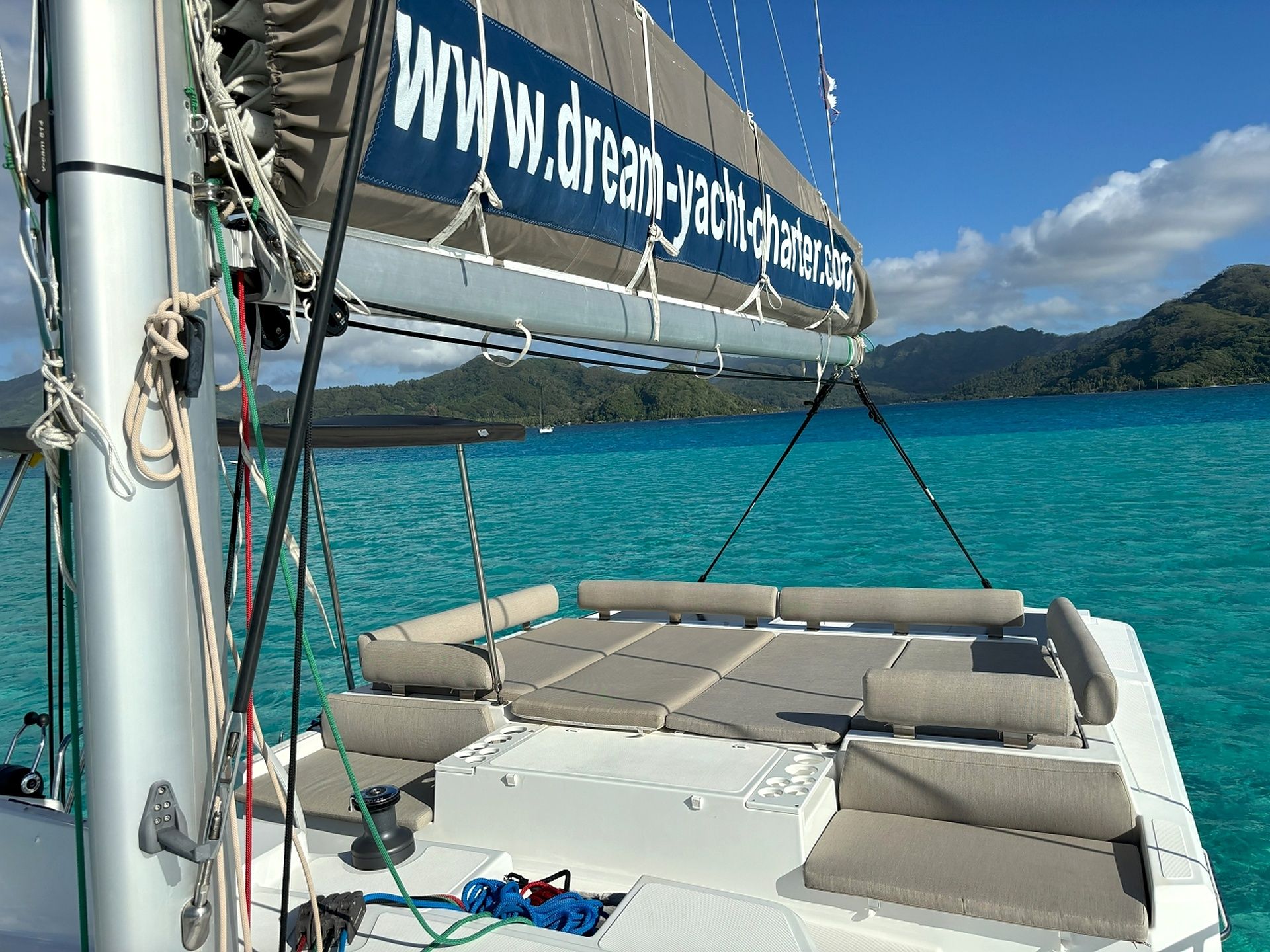 A sailboat with a website banner, a cabin with cushioned seating, and a view of a turquoise sea and distant mountains.