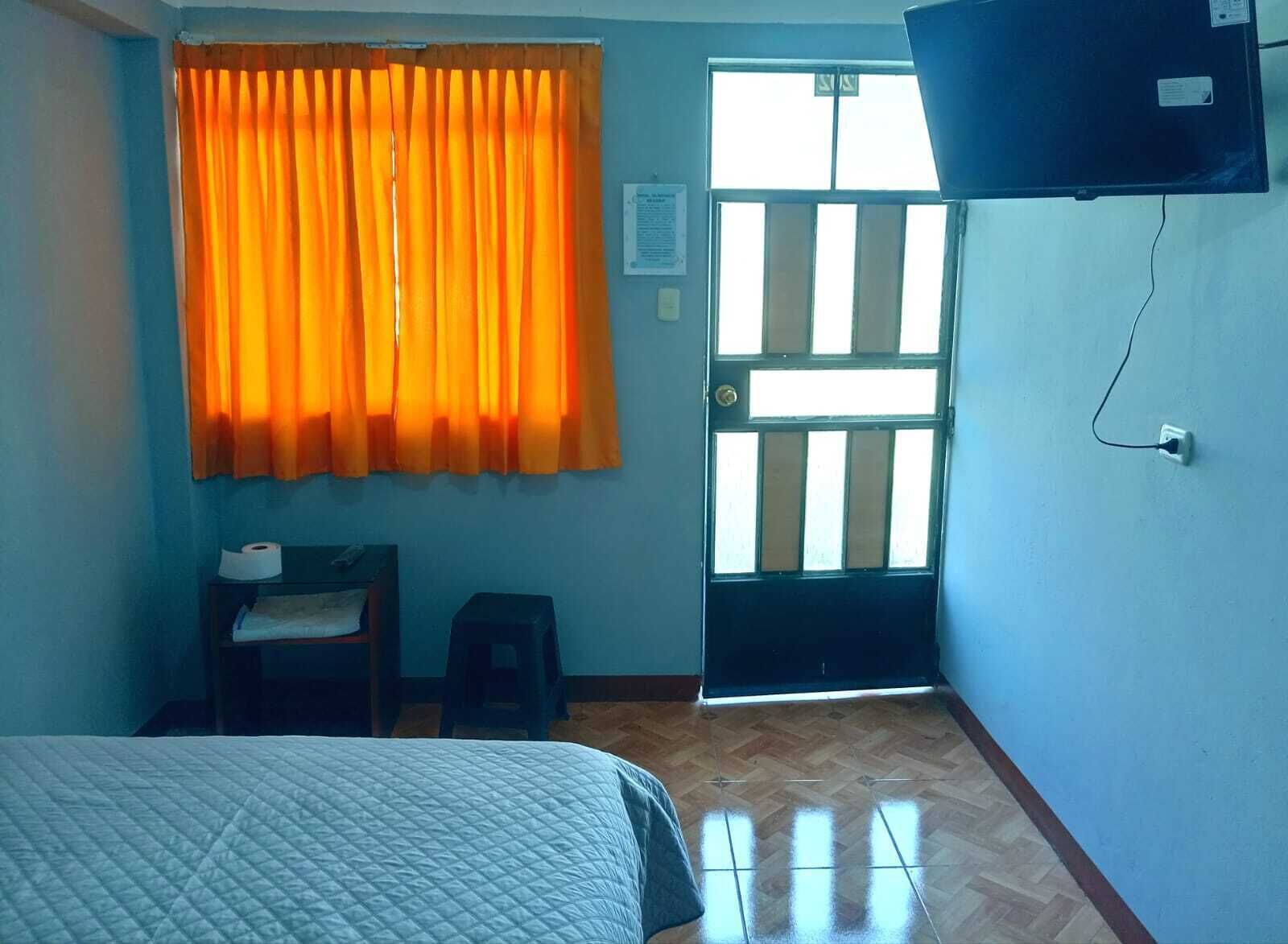 A room with a bed, a desk with a chair, a window with orange curtains, and a door with a window.