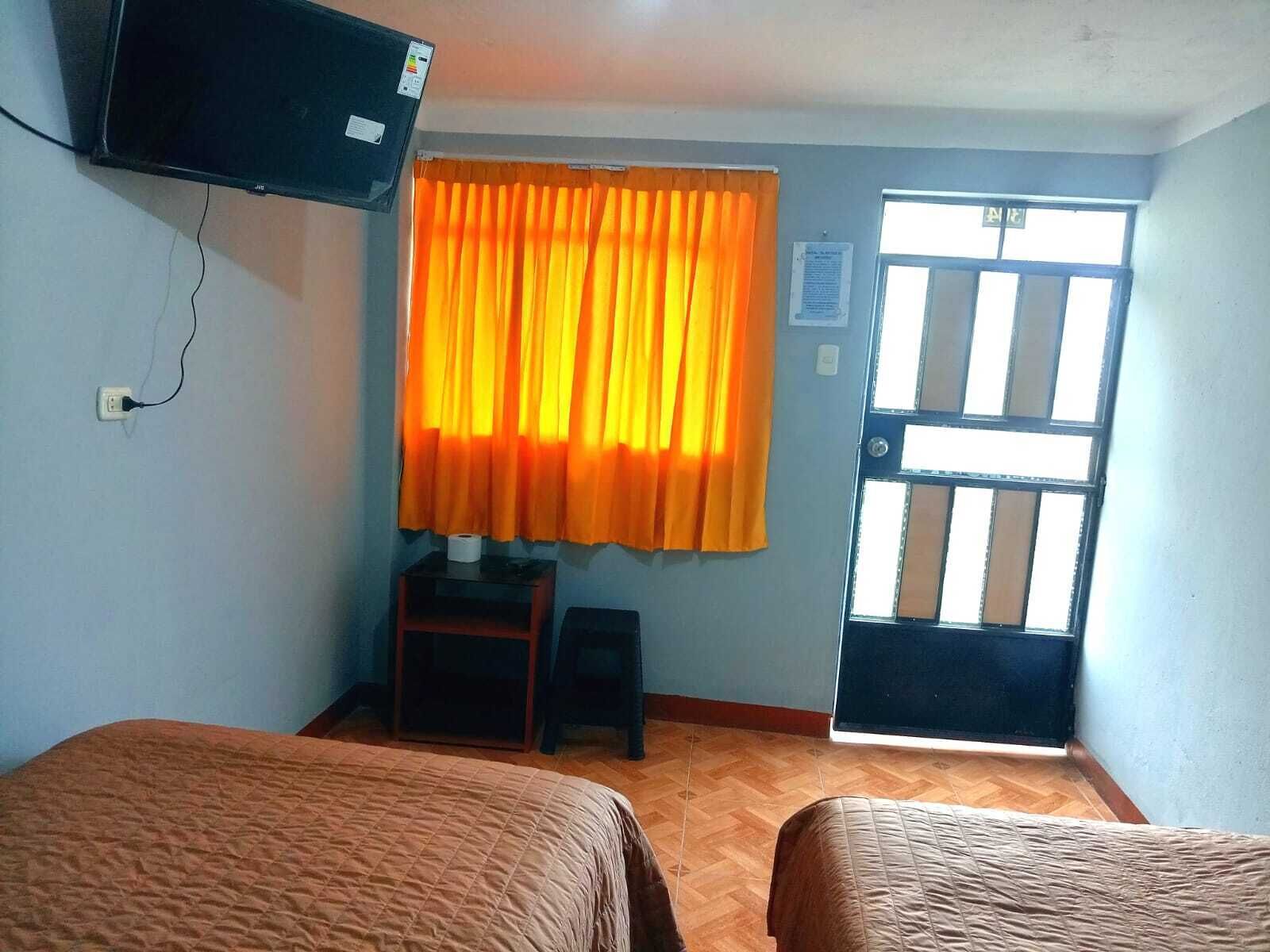 Una habitación con dos camas, un televisor montado en la pared, una ventana con cortinas naranjas y una puerta con un ventanal con barrotes.
