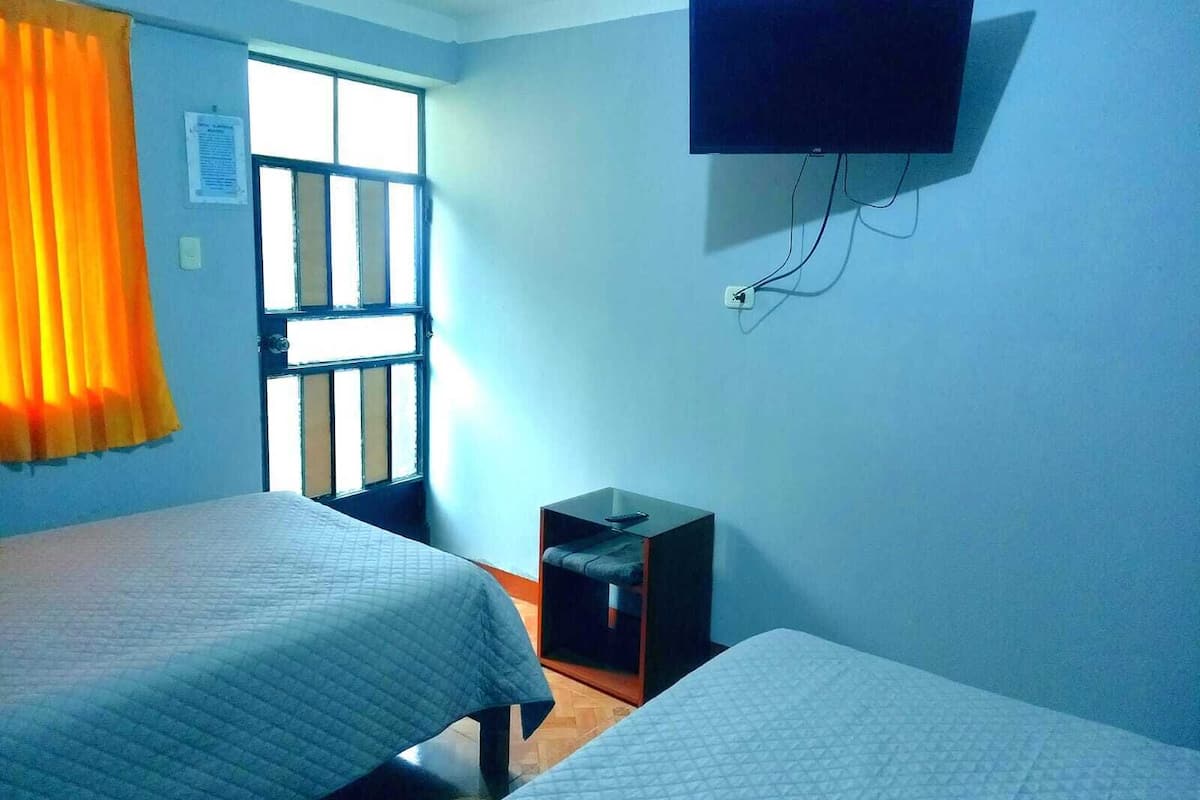 Una habitación con dos camas, un televisor montado en la pared y una ventana con cortinas.