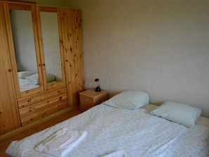 A bedroom with a wooden wardrobe, a bedside table with a lamp, and a bed with pillows and a folded towel.