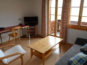 A room with a wooden table, two chairs, a sofa, a television, and a window with curtains.