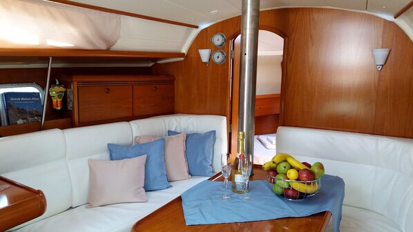 A boat cabin with a white cushioned bench, a table with a fruit bowl and glasses, and a book titled 'Greek Waters Pilot' on the wall.