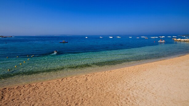 Uma praia com águas cristalinas, areia dourada e barcos ao longe.