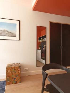 Ein Zimmer mit Holzboden, einem Esstisch mit Stühlen, einem Holzwürfel, einem gerahmten Bild an der Wand und einer durchgehenden Tür erkennbaren Küche.