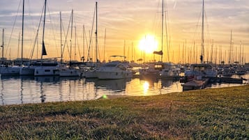 Un puerto deportivo con barcos amarrados, un primer plano con césped y el sol poniéndose al fondo.
