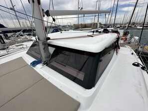 A sailboat deck with a cushioned seating area, a mast, and a view of other boats in the marina.