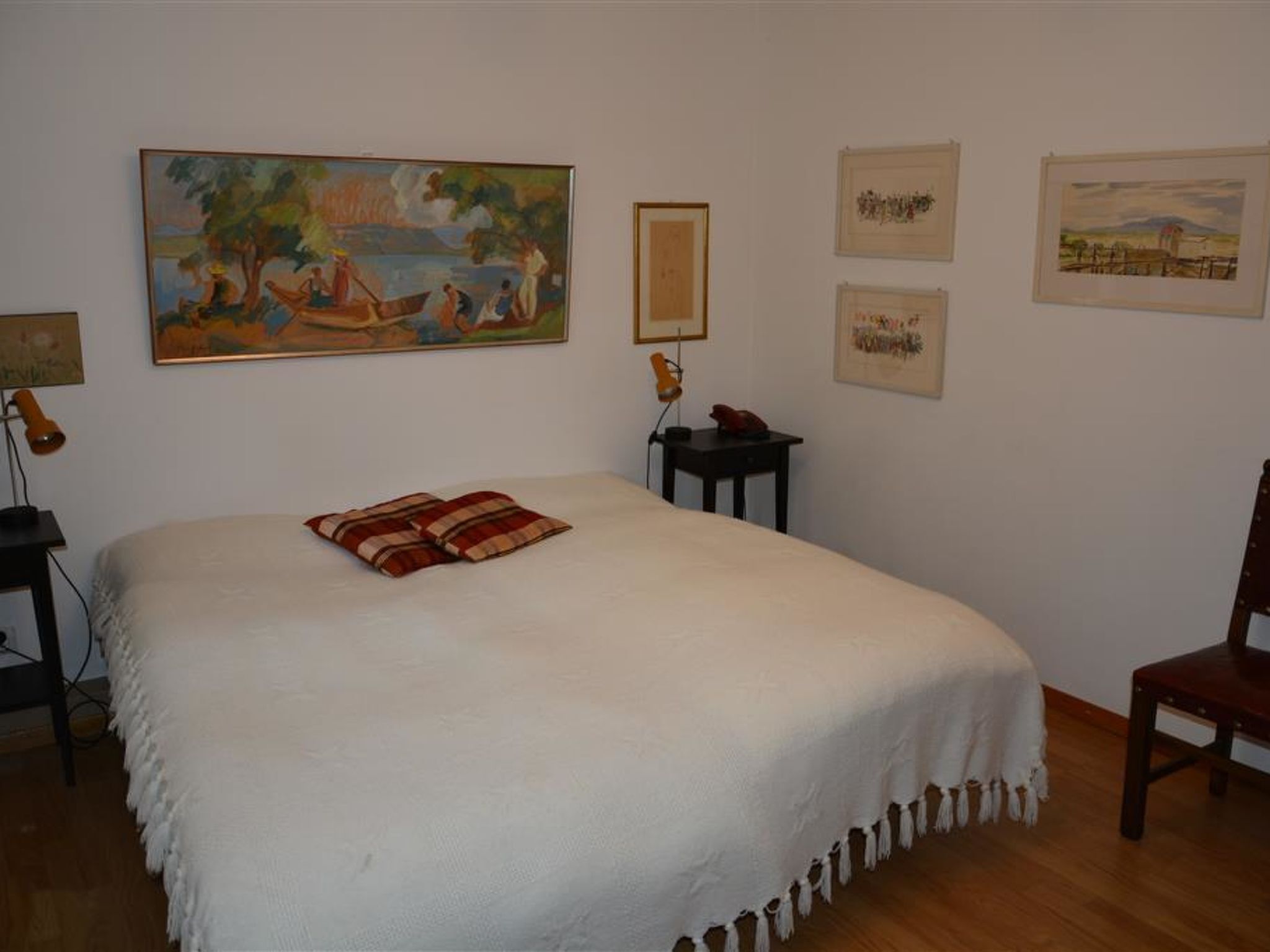 Een slaapkamer met een groot bed, een nachtkastje, een bureau en een stoel. Er hangen vier kunstwerken in lijsten aan de muur.