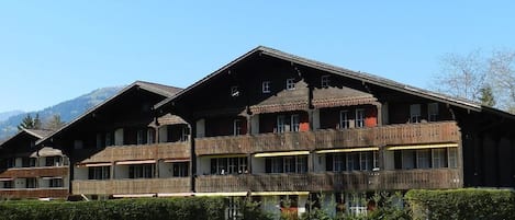 A traditional chalet-style building with multiple balconies, a gravel driveway, and a mountainous backdrop.