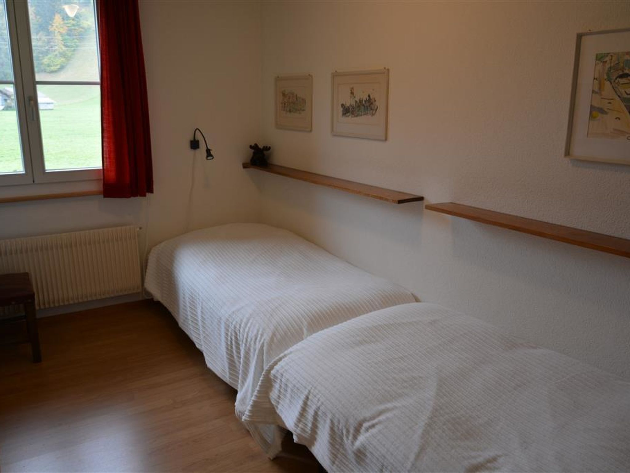 A bedroom with two single beds, a radiator, a shelf with pictures, and a window with curtains.