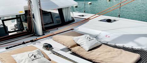 A marina with boats docked, a white yacht with beige cushions and pillows, and a forested hill in the background.