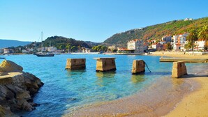 A coastal scene with clear water, a sandy beach, and buildings in the background.