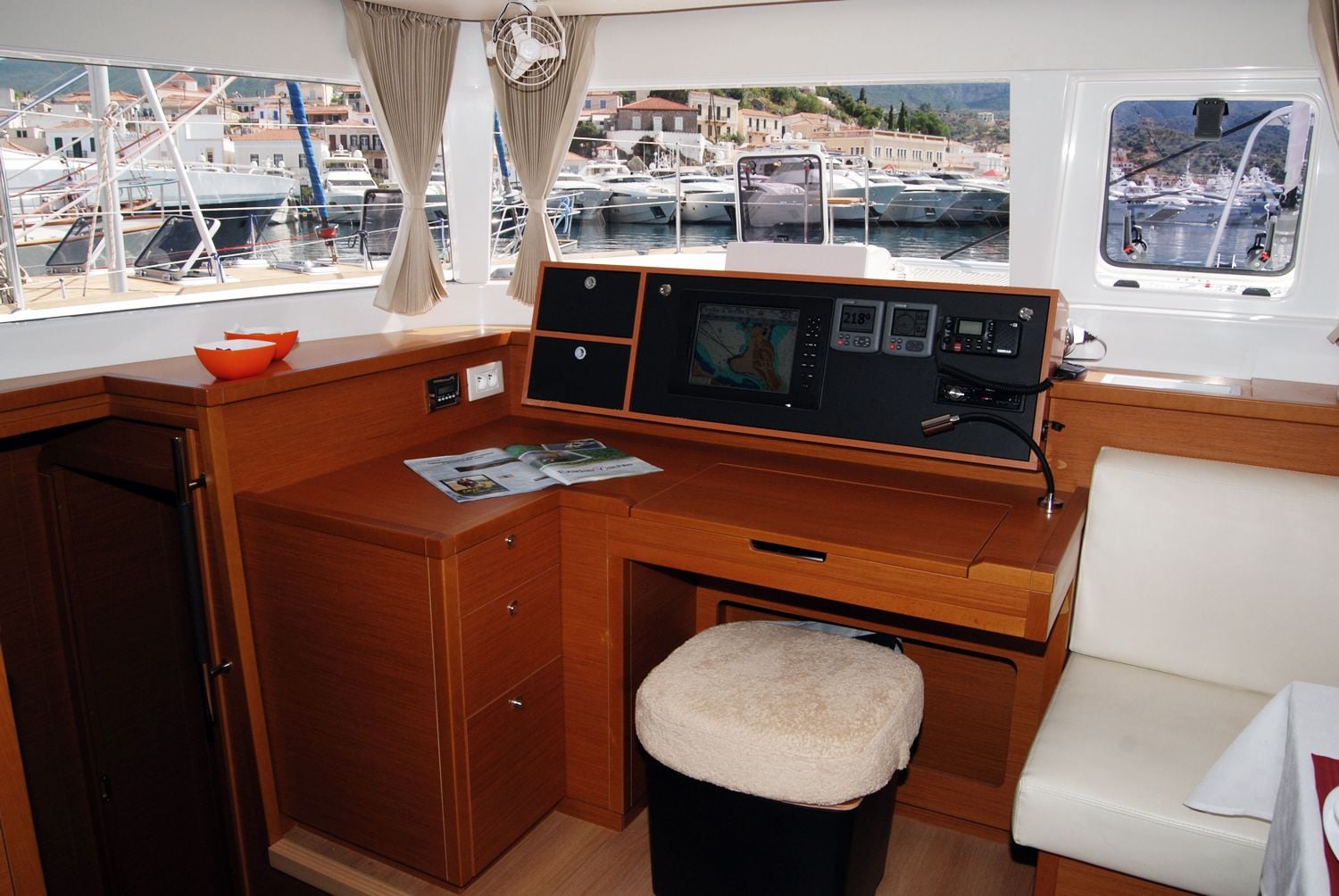 A cabin with a wooden desk, a cushioned stool, a control panel, and a view of a marina.