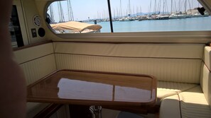 Schiffsinnenraum mit gepolsterten Sitzen, einem Holztisch und Blick auf eine Marina mit mehreren Segelbooten.