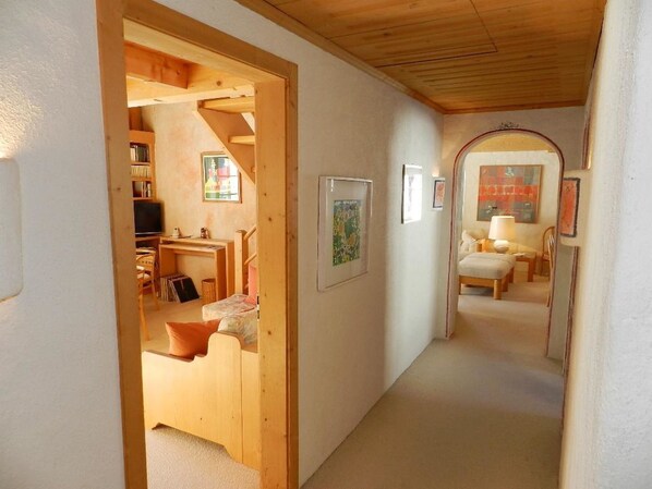 A narrow hallway with a wooden door leading to a bedroom with a bed and a desk. There are framed pictures on the walls and a lamp on a side table.