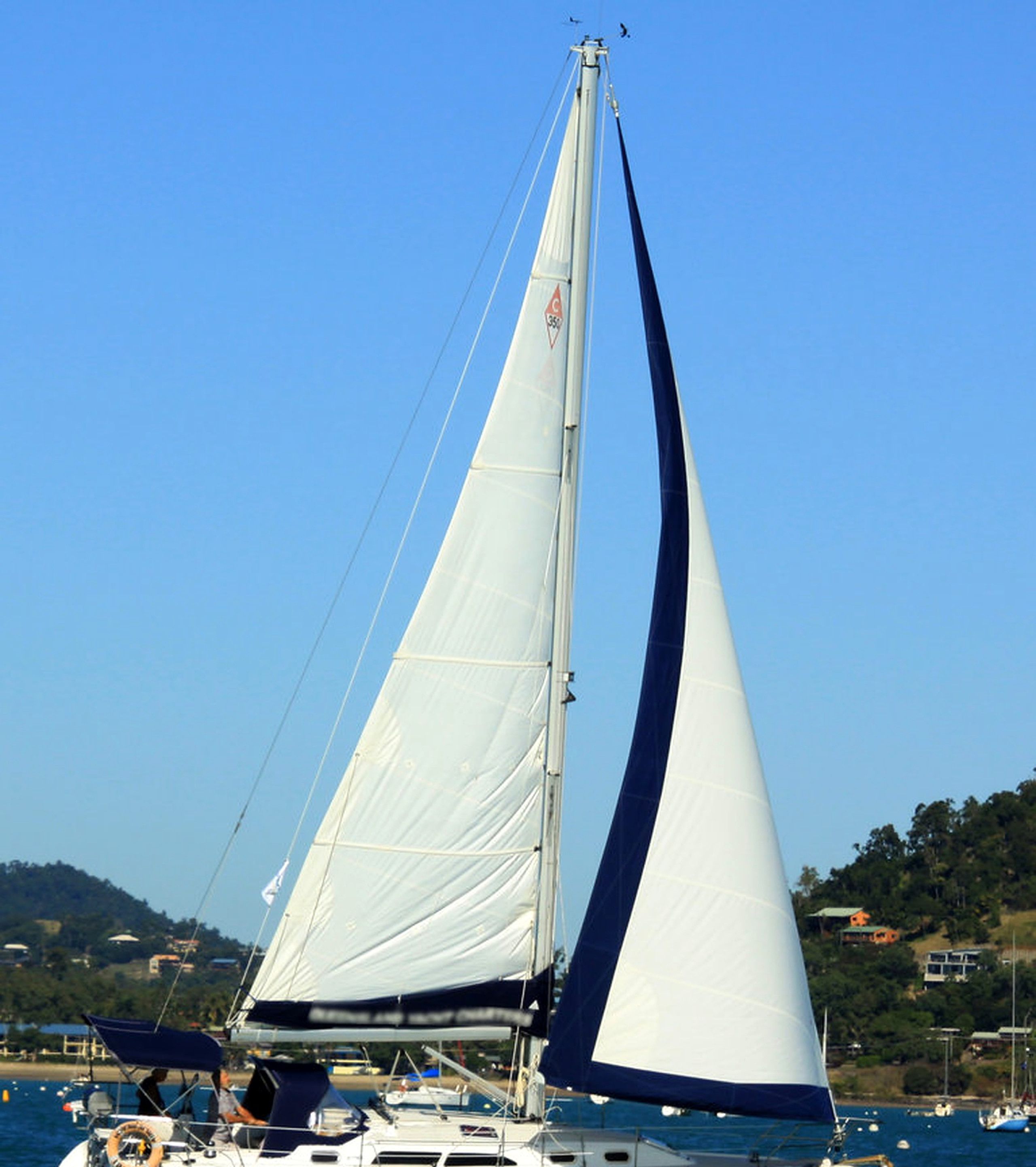 Ein Segelboot mit hohem Mast und zwei Segeln liegt in einer Marina vor Anker, im Hintergrund eine hügelige Landschaft und weitere Boote.