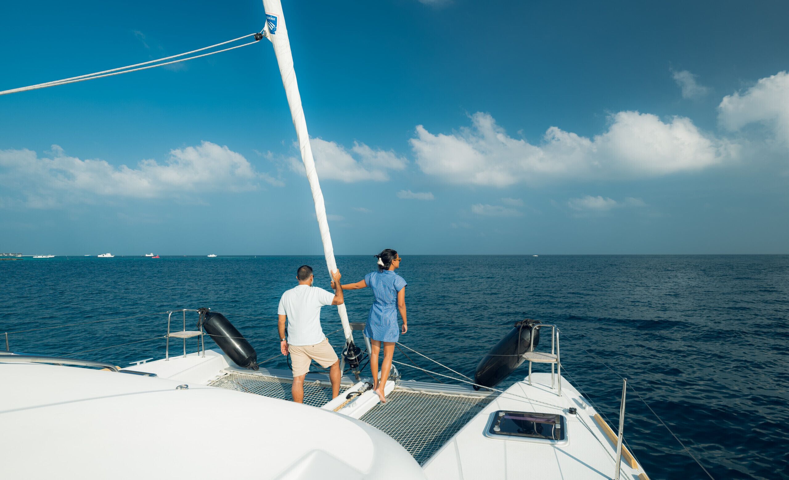Un velero con dos personas de pie en la proa, cielo despejado y mar tranquilo.