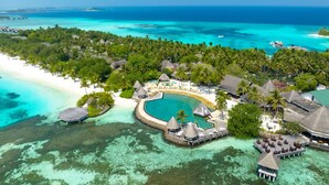 Vista aérea de un resort tropical con una piscina grande, bungalows sobre el agua y una playa.