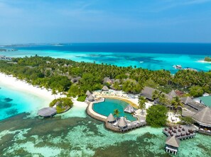An aerial view of a tropical resort with a large pool, overwater bungalows, and a beach.