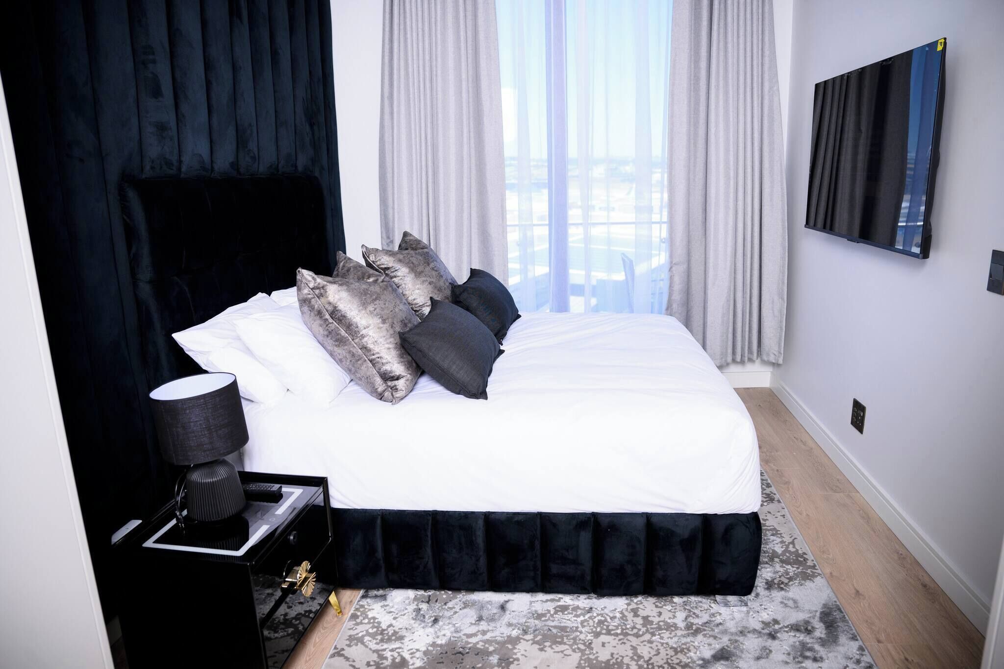 A modern hotel room with a large bed, a black nightstand, a lamp, and a view of the city through large windows.