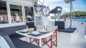 A dining area with a table set for two, a cushioned bench, and a view of the sea and hills. - Sun-kissed Sailing Adventure in the Ionian Islands (Preveza)