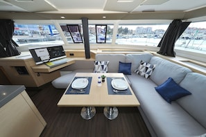 A boat interior with a dining area, a sofa, a desk with a computer, and a view of a marina.