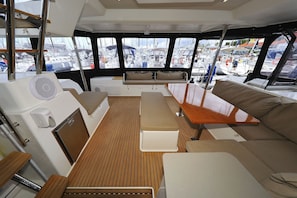 A boat interior with a wooden deck, cushioned seating, and a view of other boats through large windows.