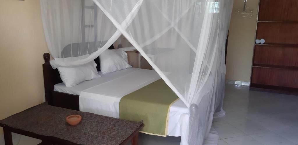 A single bed with a canopy, a small table with a bowl, and a wooden wardrobe in the background.