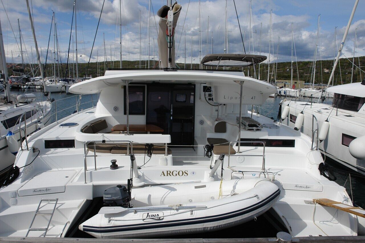 Un yate llamado ARGOS atracado en un puerto deportivo con varias embarcaciones, una cabina con zona de estar y un mástil visible.