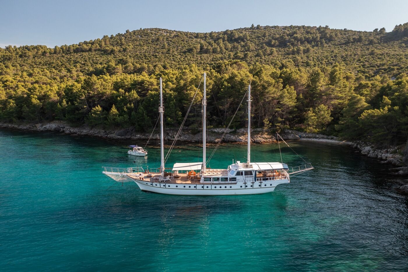 Eine weiße Yacht mit mehreren Masten und einer überdachten Deck, vor Anker in türkisfarbenem Wasser, mit einem bewaldeten Hügel im Hintergrund.