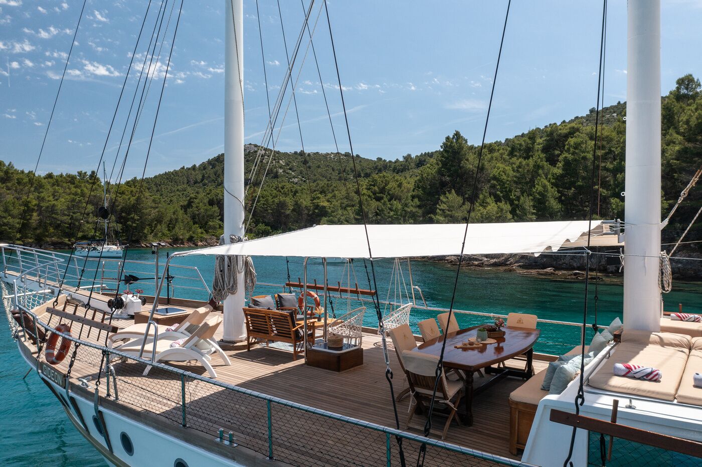 A sailboat with a deck area featuring outdoor seating, a white canopy, and a view of a forested coastline.