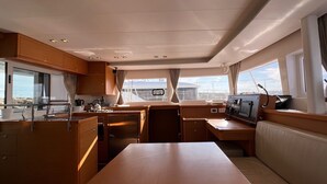 Interni di uno yacht con un tavolo da pranzo centrale, mobili in legno, un pannello di controllo e ampie finestre vista mare.