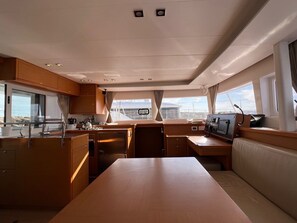 A yacht interior with a central dining table, wooden cabinetry, a control panel, and large windows offering views of the sea.