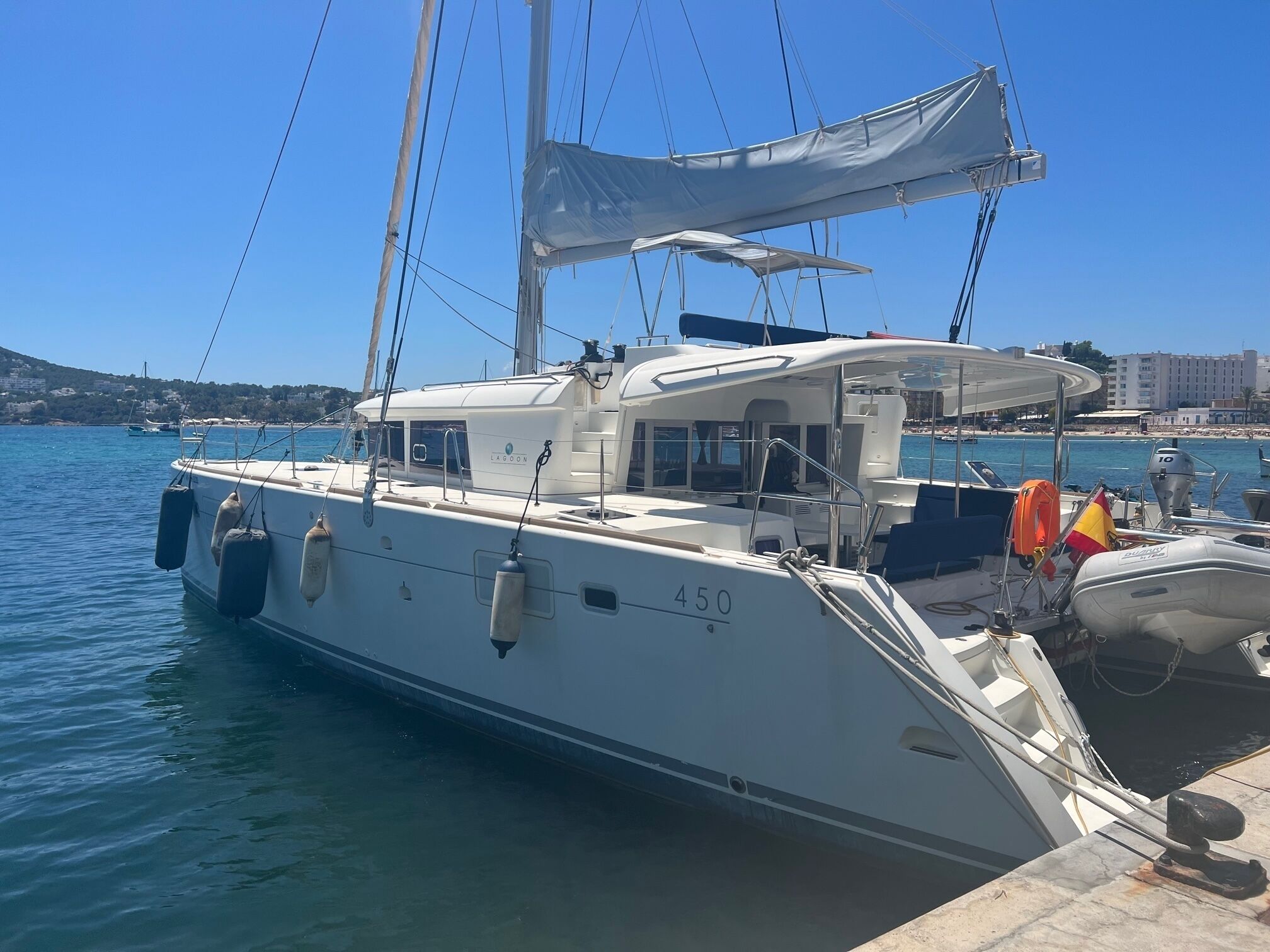 Eine Segelyacht liegt in einer Marina vor Anker, der Himmel ist wolkenlos, das Wasser ruhig und in der Ferne ist eine Küste mit Gebäuden zu sehen.