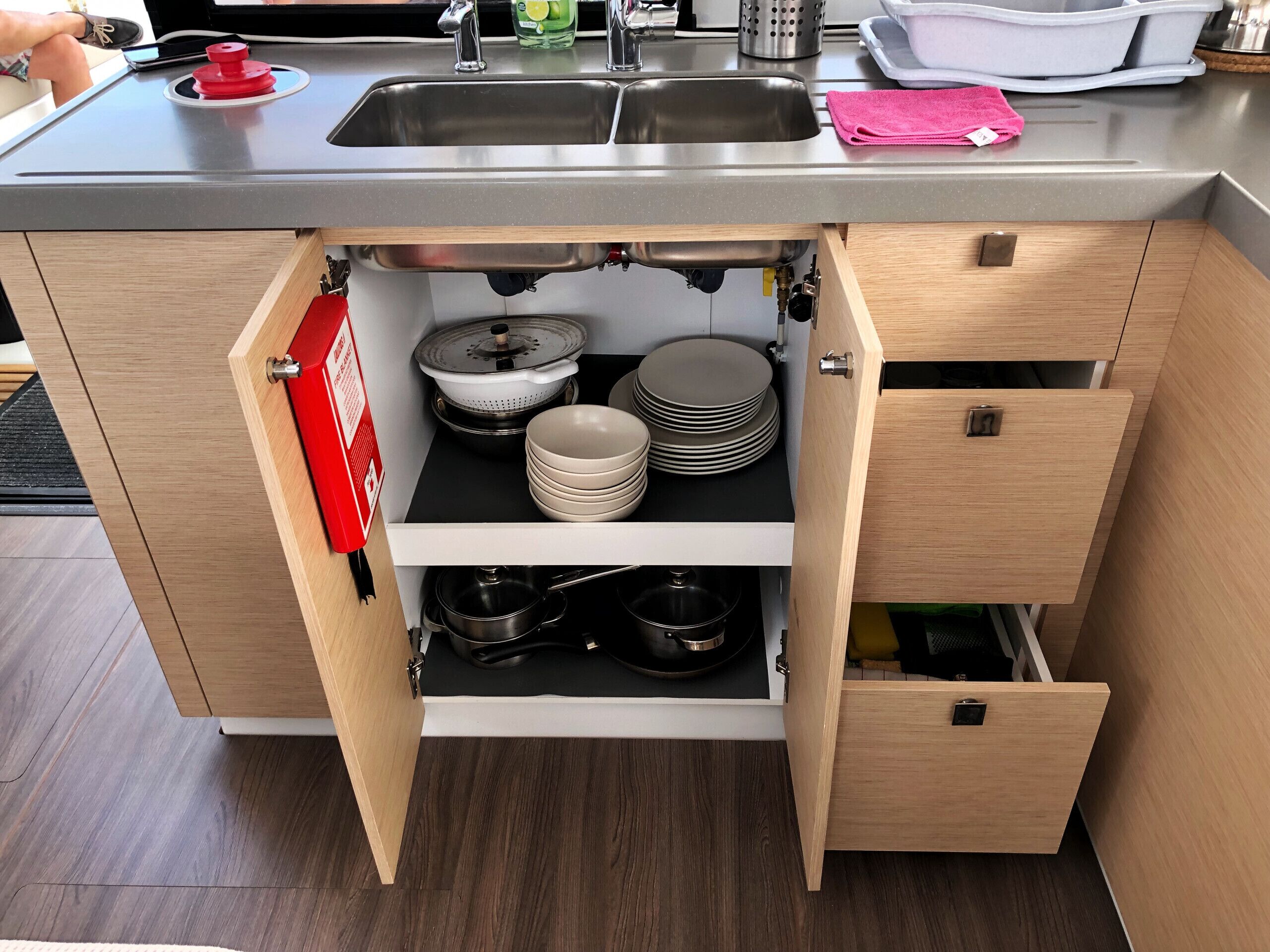 A kitchen with a stainless steel sink, wooden cabinets, and a built-in drawer unit with pots and plates.
