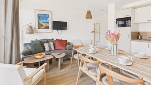 Un salon moderne comprenant un canapé, une table à manger avec des chaises, un téléviseur fixé au mur et un coin cuisine avec des placards et des appareils électroménagers.