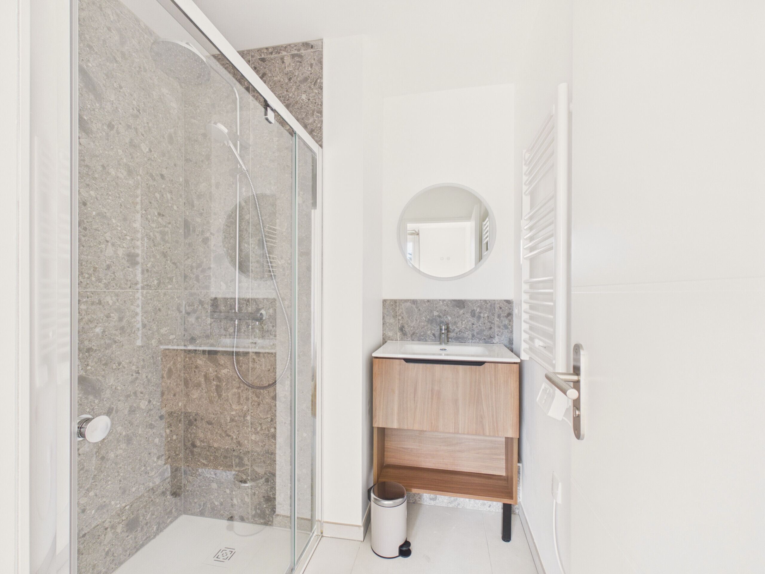 A modern bathroom with a glass-enclosed shower, a wooden vanity, and a round mirror.